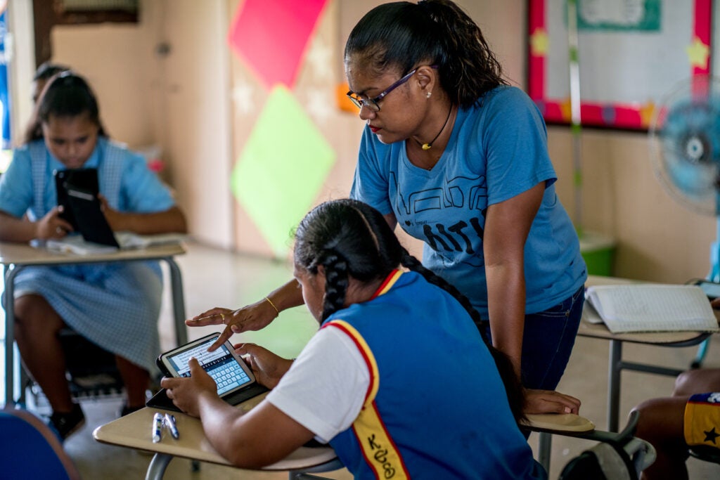 A girl is sitting at a desk and typing on a tablet screen in a school classroom. A woman is leaning over to assist her and point out something on the tablet.